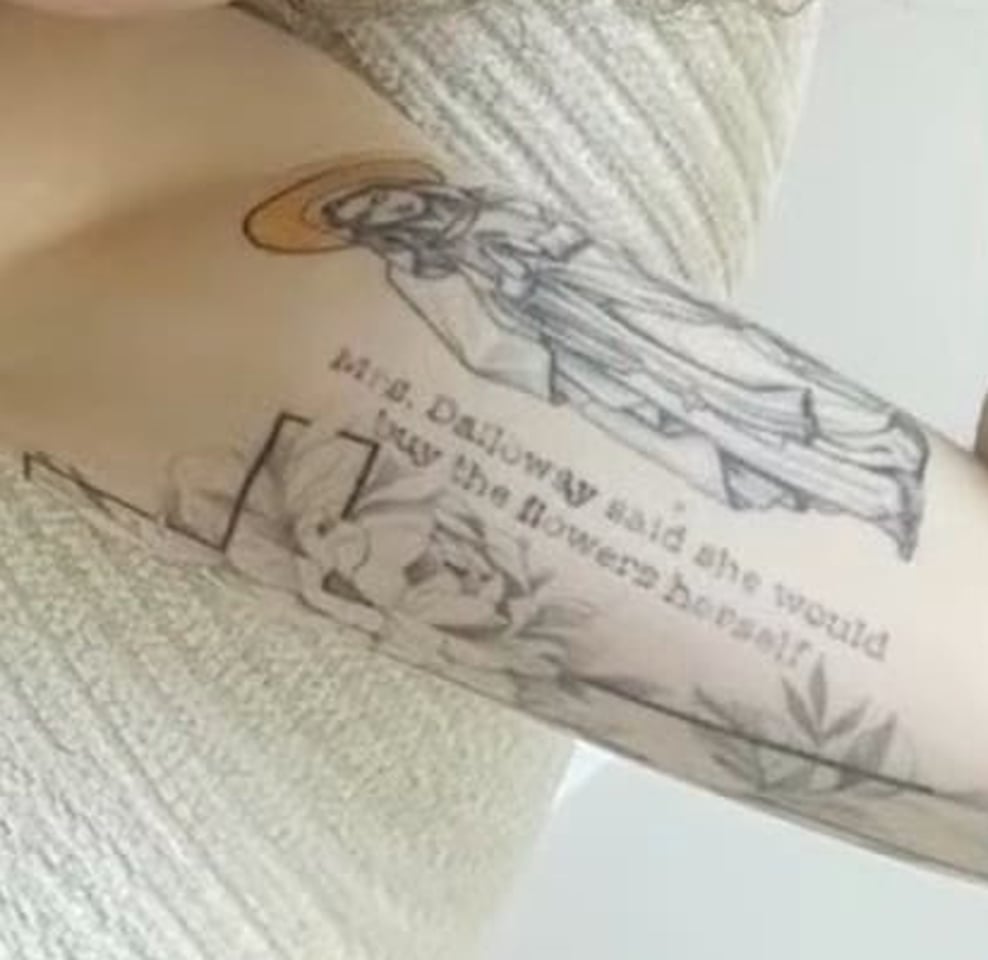 Close-up of Nancy Mace’s left arm: a robed saint figure with a gold halo above the fine-line script "Mrs. Dalloway said she would buy the flowers herself," beside a bold, rose-covered squared structure (a cross or a small arch), the saint's robe draping down alongside it.