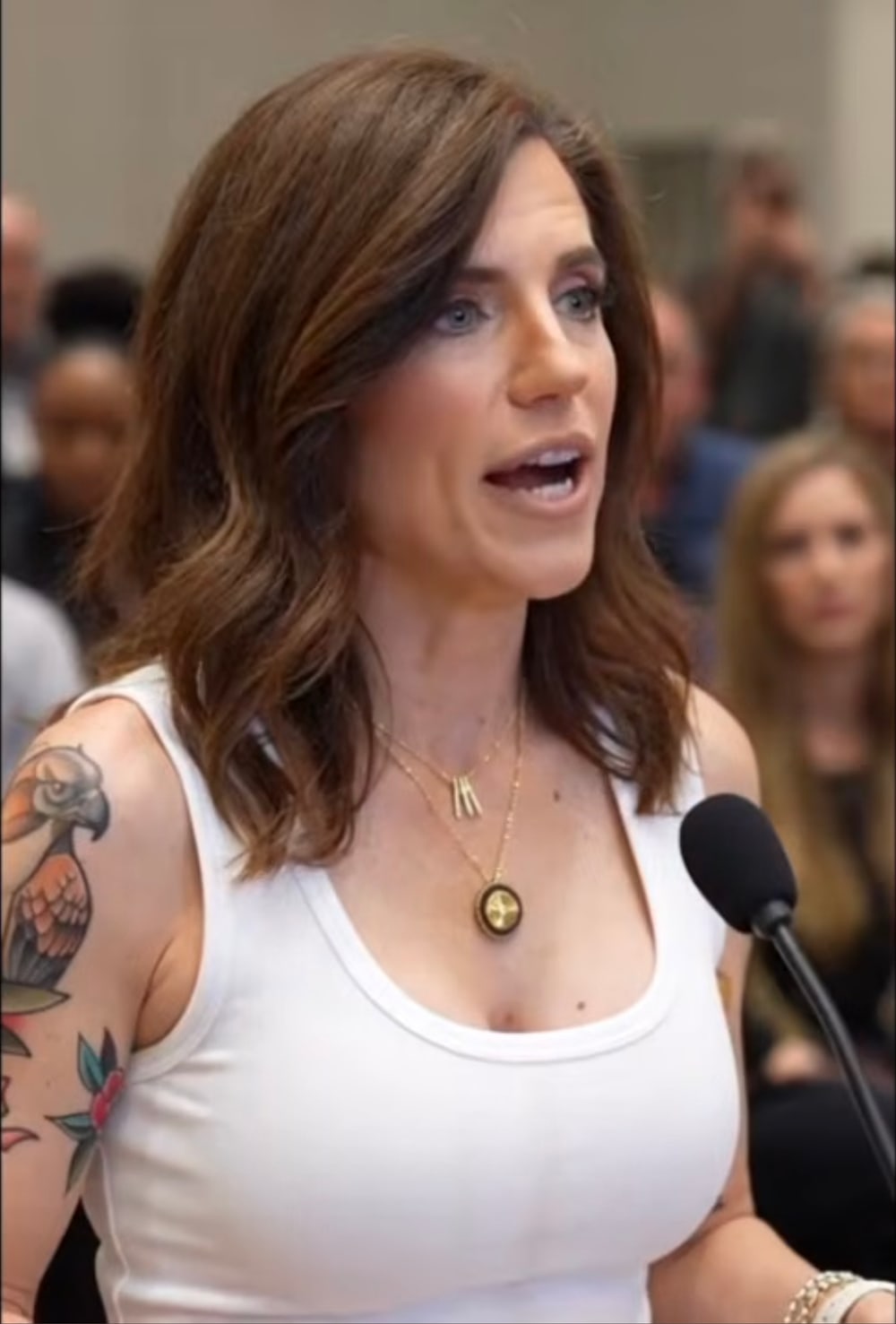 Microphone frame from the GET MACED intro: a woman identifiable as Nancy Mace in a white tank top speaking at a microphone before a crowd, with a colorful American-traditional bird across her upper left arm and a rose below it.
