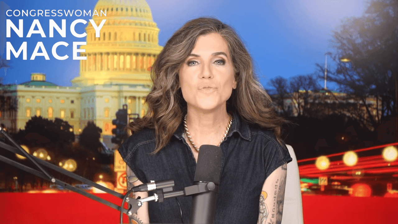 A second studio-desk frame from the GET MACED intro: Nancy Mace at the microphone with the CONGRESSWOMAN NANCY MACE lower-third, the same forearm script visible.