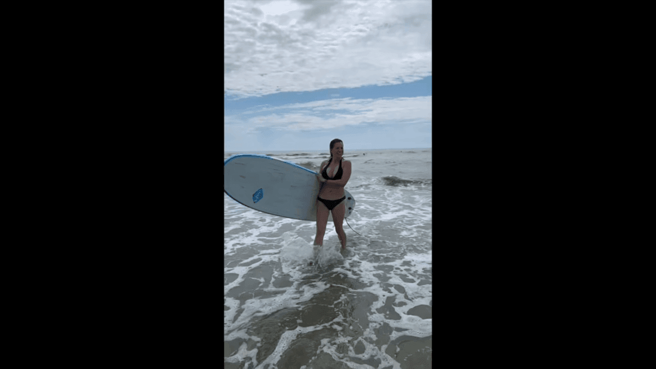 Frame from Nancy Mace's own Get Maced footage: she carries a surfboard out of the water in a swimsuit, her arms visible.
